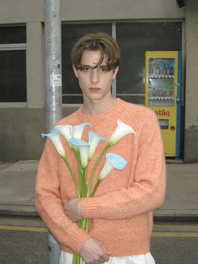 Medium shot of a young person with a deadpan face, clutching pale blue calla lilies to their chest. Standing on a worn g