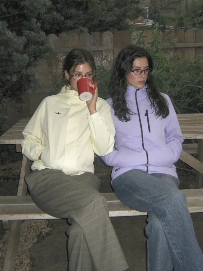Two young adults sitting side by side on a wooden bench at a worn picnic table in a small, plant-filled backyard, both w